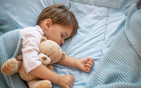 کودک آرام خوابیده در تخت با نور ملایم چراغ خواب و عروسک دلبستگی